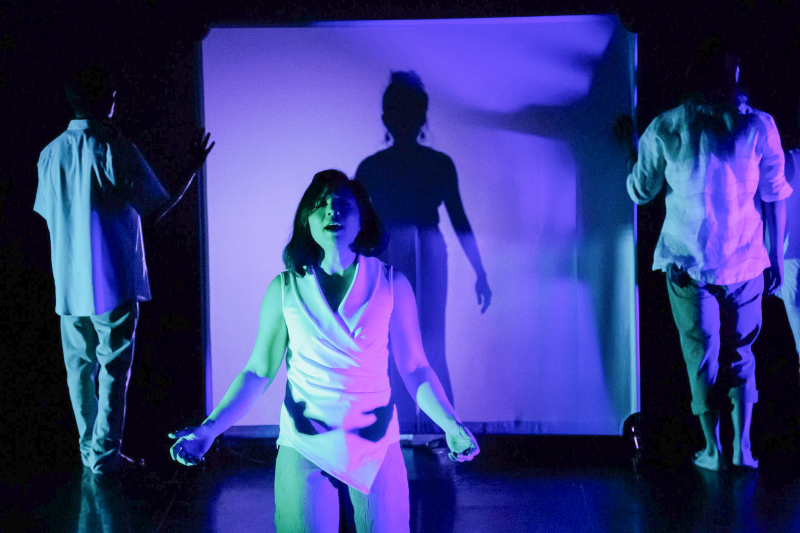 A performance photo in purple/blue light. Megan Lowe sings and stands center stage with dancers in the background.