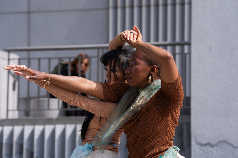 Two dancers connect with outstretched arms touching and the other hand above their heads, clasped together. 